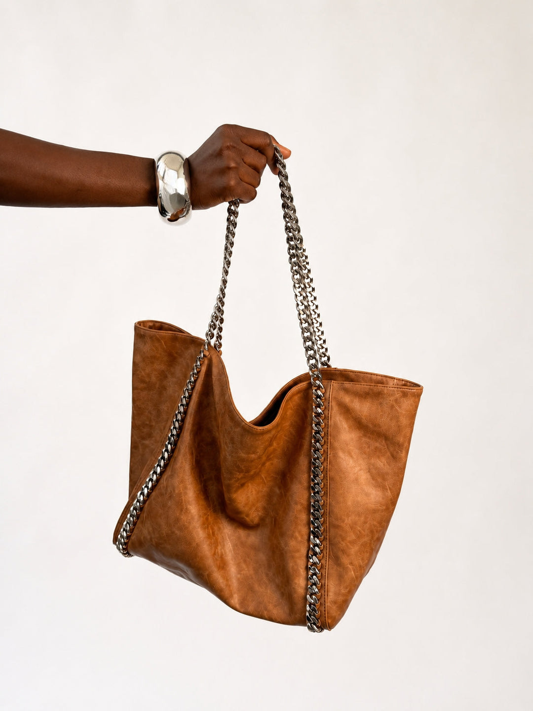 The Chain Tote in Cognac — side view of slouchy faux leather tote with silver curb chain straps against neutral backdrop