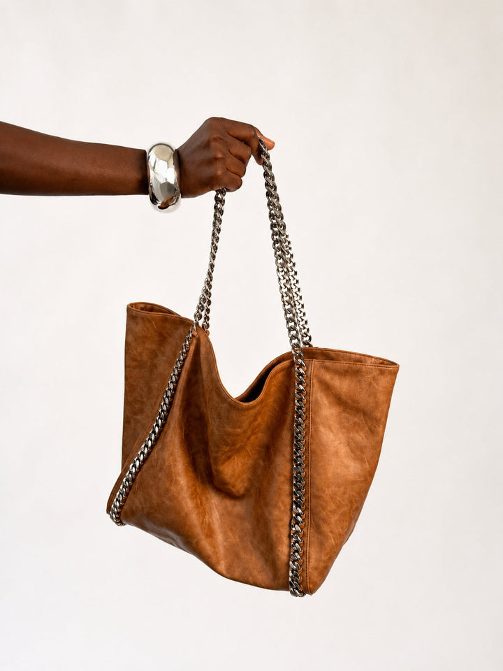 The Chain Tote in Cognac — side view of slouchy faux leather tote with silver curb chain straps against neutral backdrop
