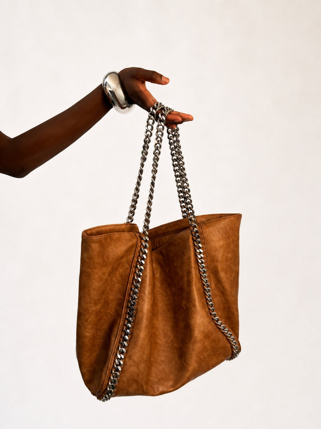 The Chain Tote in Cognac — oversized faux leather tote with silver chain-link straps and chain trim, held up by model