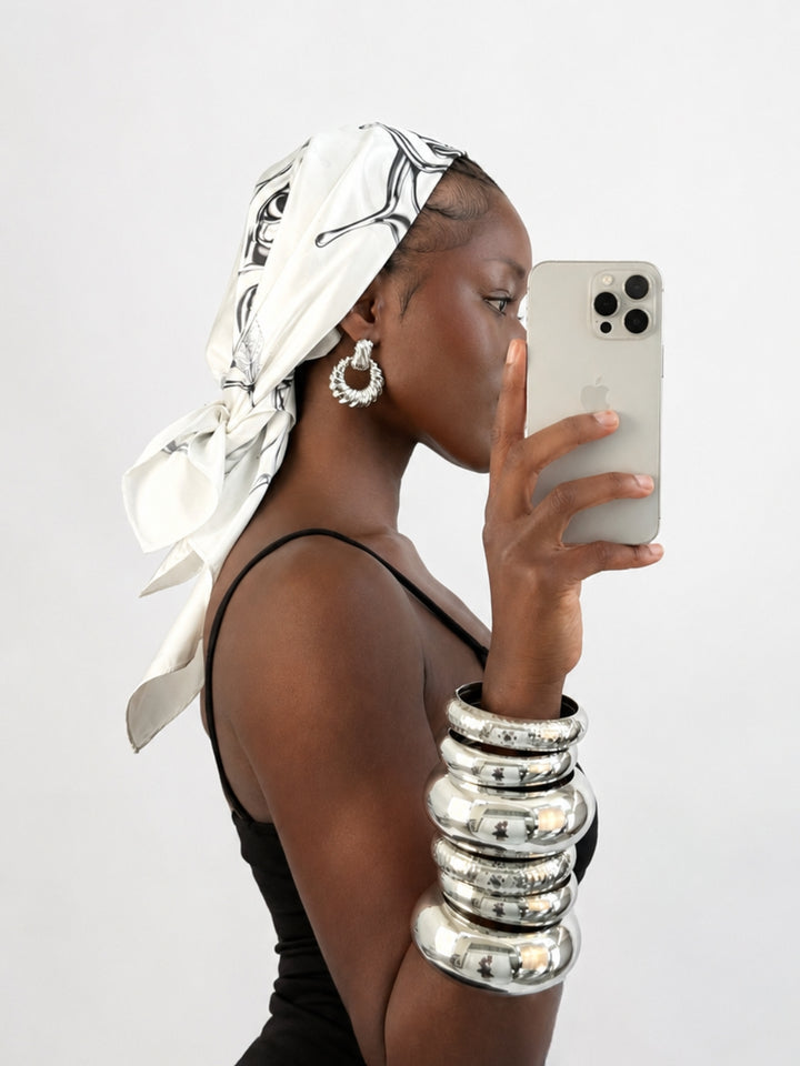 Woman taking a selfie with a phone, wearing a headscarf and bracelets, on a plain background
